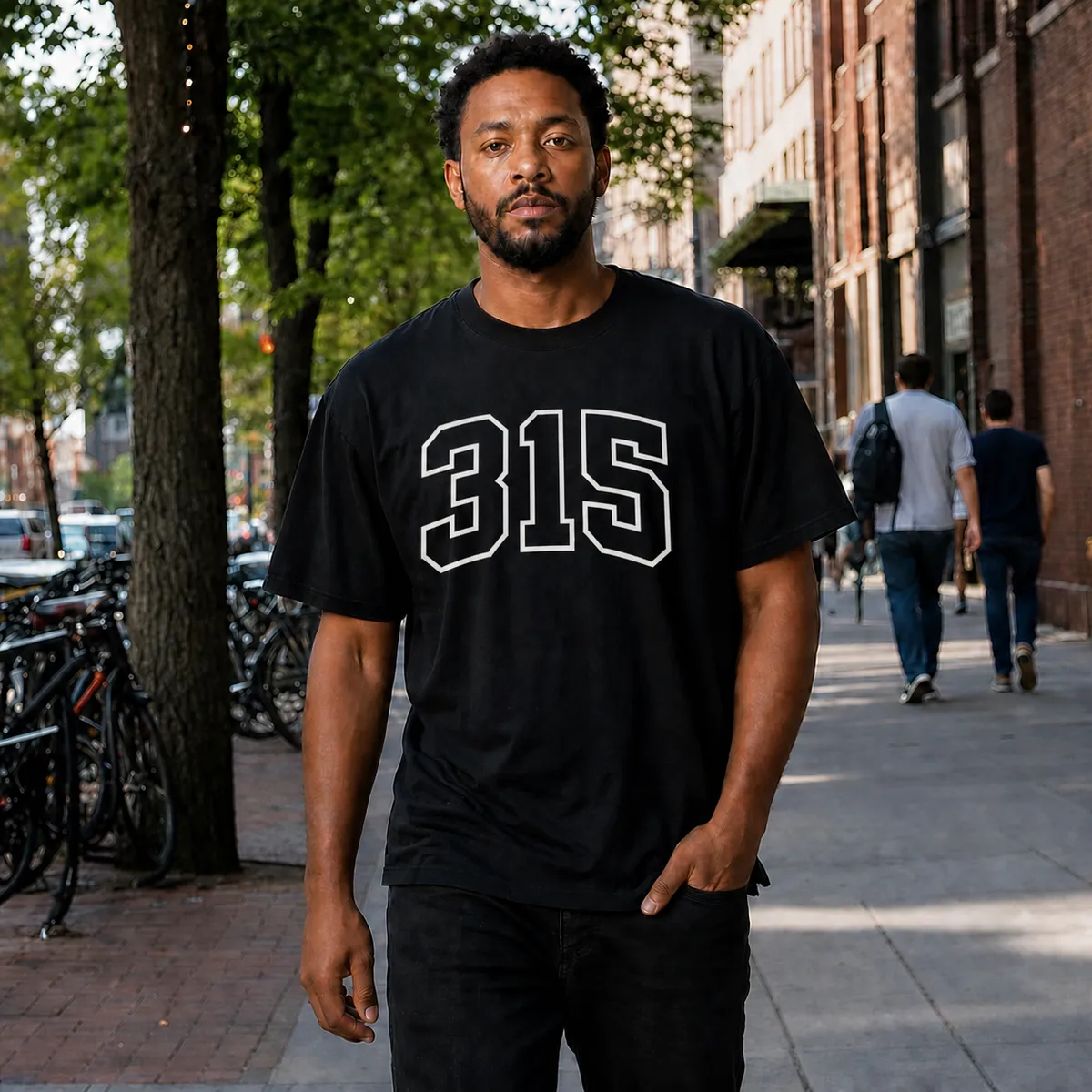 A man on a tree-lined city sidewalk wearing the black 315 Varsity tee, the area code printed across the chest in white outlined varsity block.