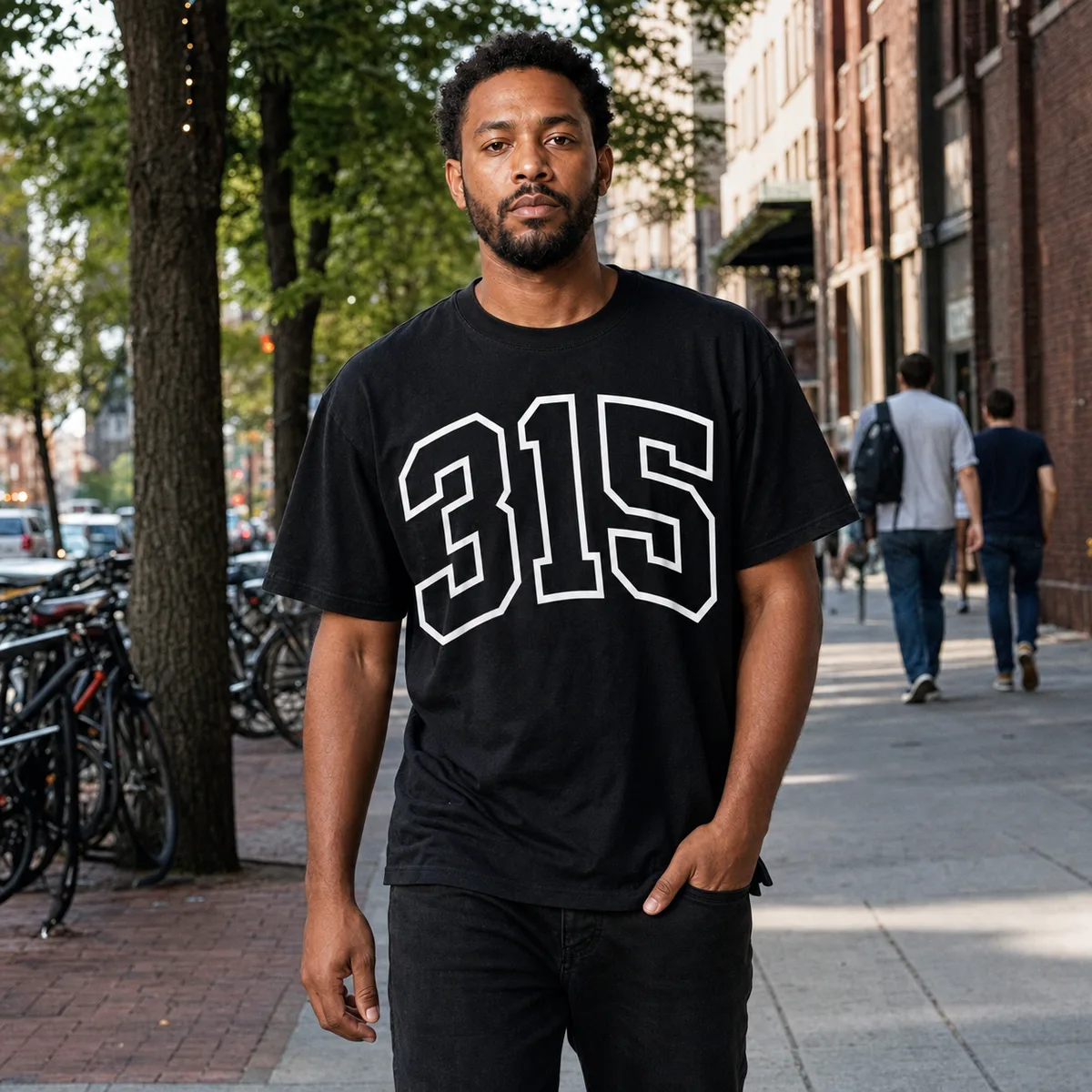 A man on a tree-lined city sidewalk wearing the black 315 Varsity tee, the area code printed across the chest in white outlined varsity block.
