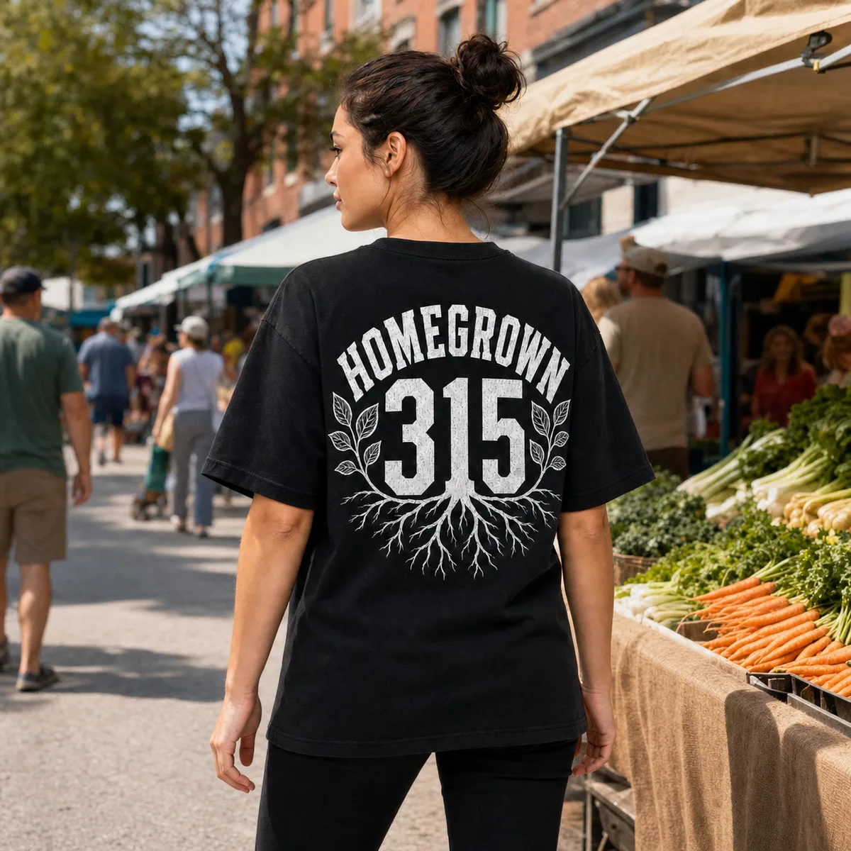 A woman walking through an outdoor farmers market, seen from behind, wearing the black Homegrown 315 tee — HOMEGROWN arched over 315 above a spreading root system, printed in white across the back.